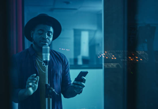 Portrait of Successful Young Black Artist, Singer, Performer Singing His Hit Song for the New Album. Wearing Stylish Hat, Holding Smartphone and Standing in Music Record Studio Soundproof Room.