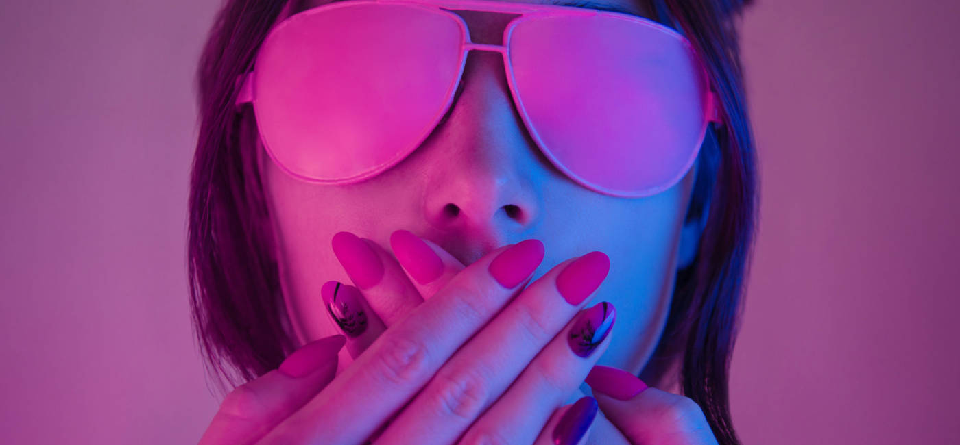 Young woman in pink glasses covers her mouth with hands with a manicure, retrowave style portrait. Demonstration of a beautiful manicure.
