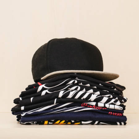 Pile-of-brand-new-t-shirts,-dark-tones,-with-print-and-a-black-straight-flap-cap-over-the-pile