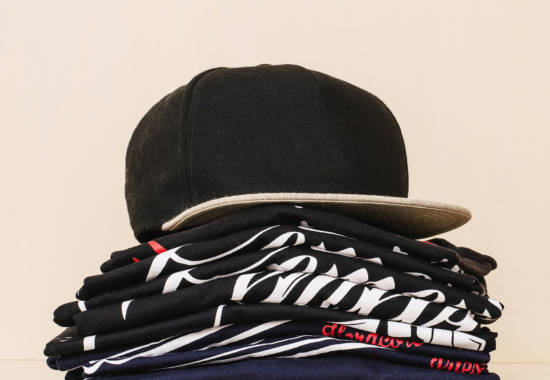 Pile-of-brand-new-t-shirts,-dark-tones,-with-print-and-a-black-straight-flap-cap-over-the-pile