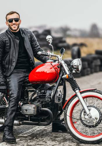 Red motorbike with rider. A man in a black leather jacket and pants stands sideways in the middle of the road. Tires are laid on the background.